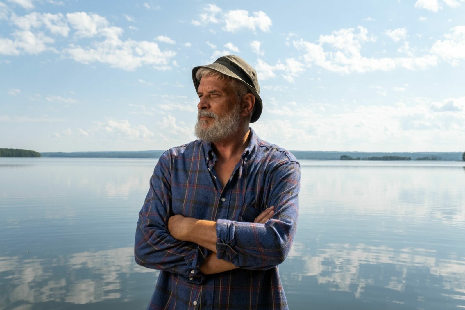 Senior man with beard and hat stands by a tranquil lake on a sunny day, arms crossed and contemplative.